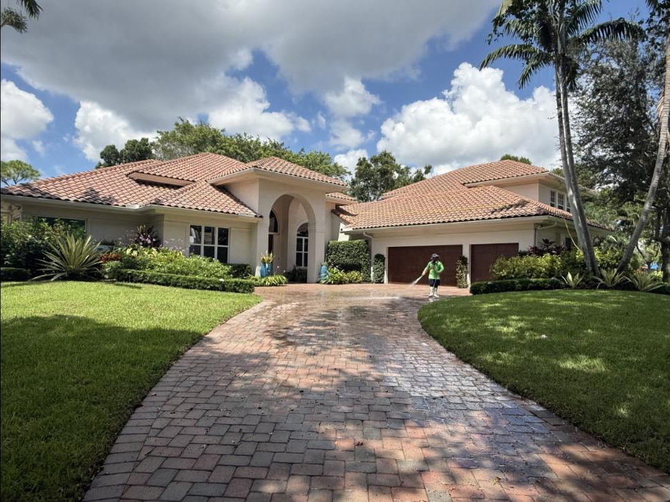 Technician rinsing a brick paver driveway at a Boca Raton estate — water arcing from the wand, barrel tile roof home behind