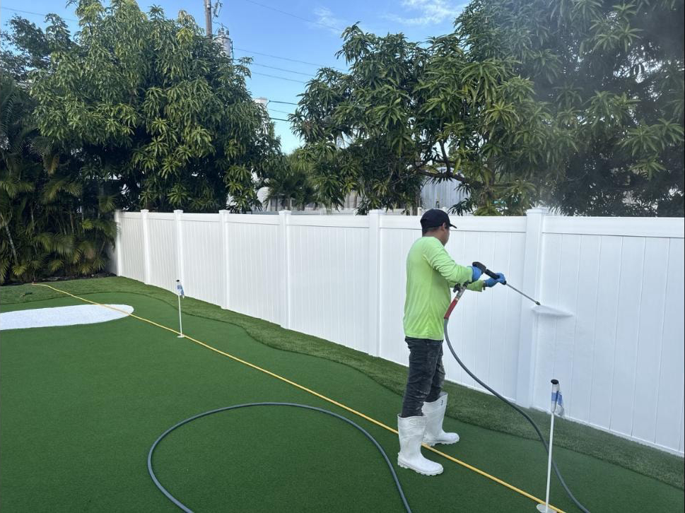 Technician soft-washing a white vinyl fence beside an artificial turf putting green — Boca Raton, FL