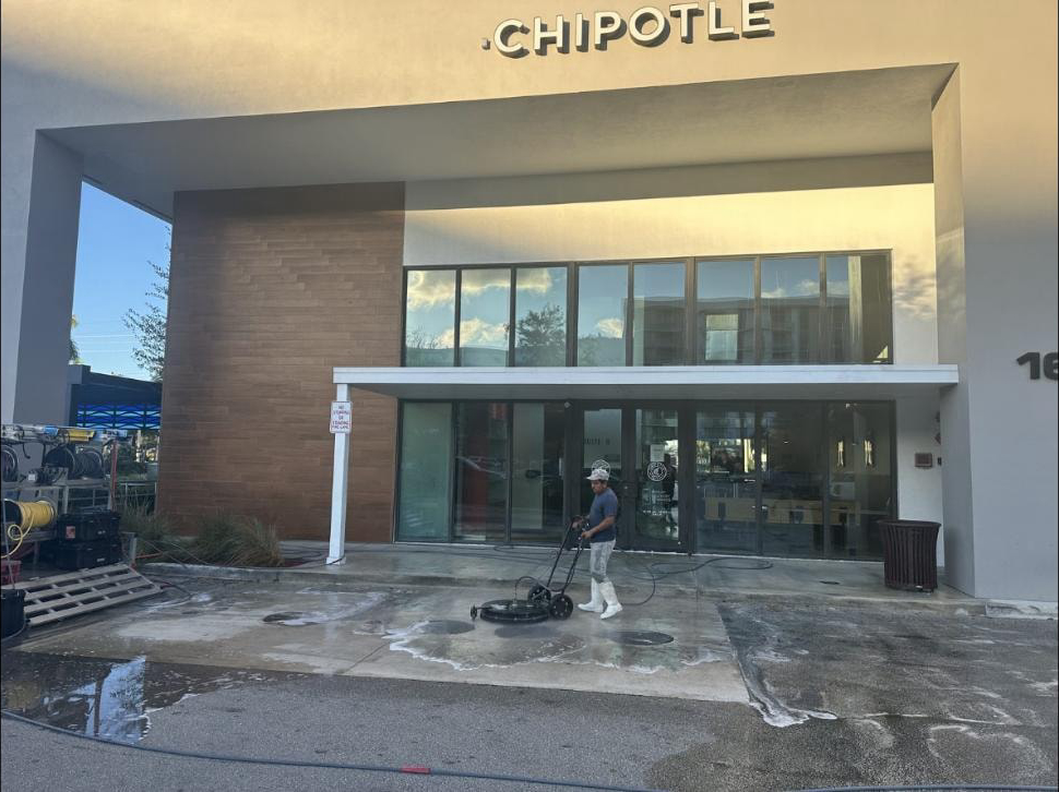 Technician running a surface cleaner at a restaurant entrance in Boynton Beach, FL — wet concrete apron, service rig at left, commercial building facade