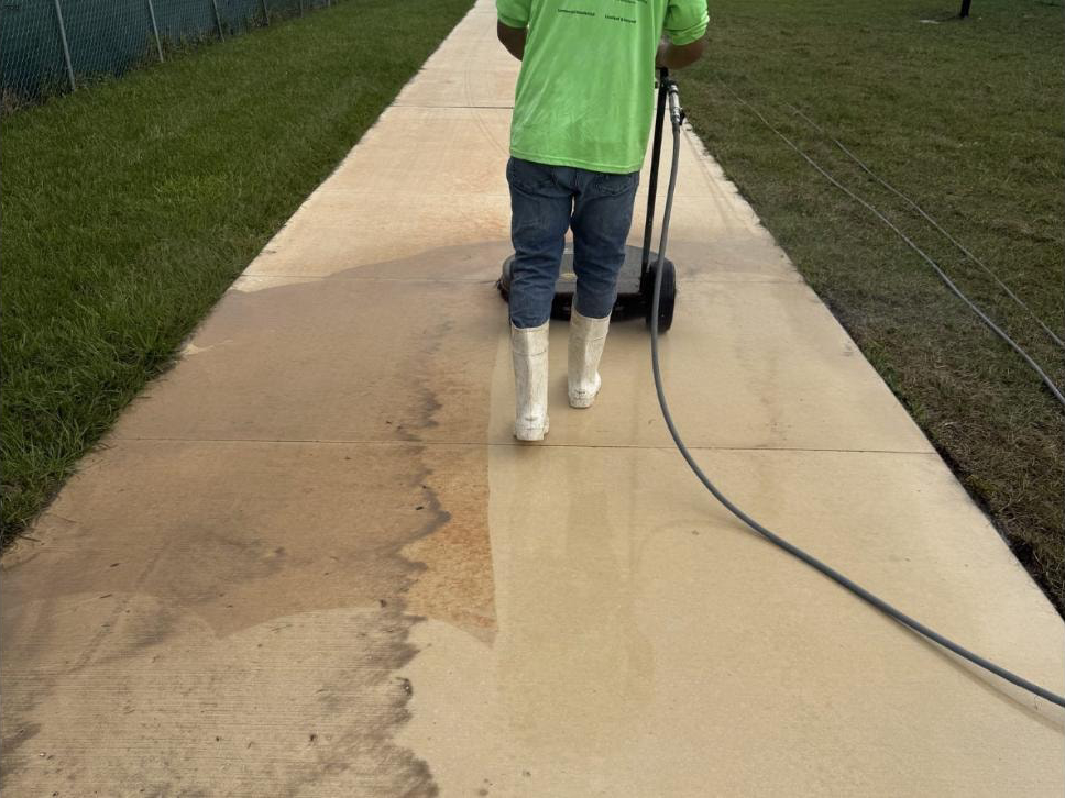 Technician running a surface cleaner on a Canyon Park concrete walkway in Boynton Beach, FL — sharp dirty-versus-clean boundary visible across the surface