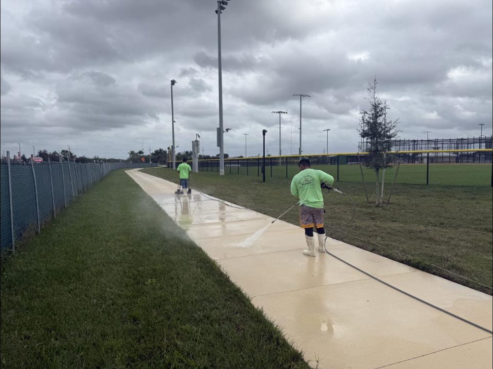 Two-man crew pressure washing a long concrete park walkway beside athletic fields at Canyon Park, Boynton Beach, FL — overcast sky, wet surface