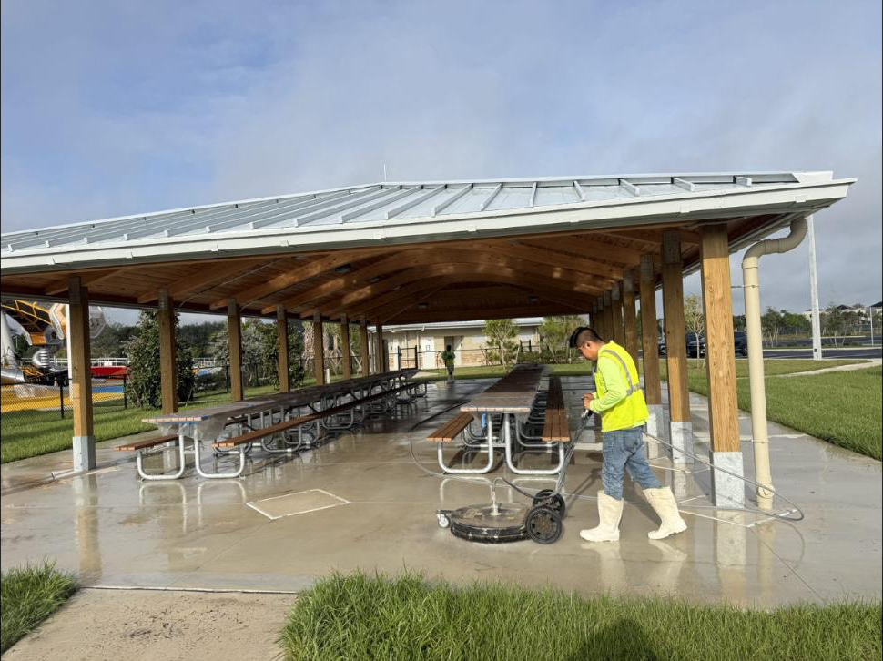 Technician running a surface cleaner under an open-air park pavilion at Canyon Park, Boynton Beach, FL — picnic tables and wet concrete floor