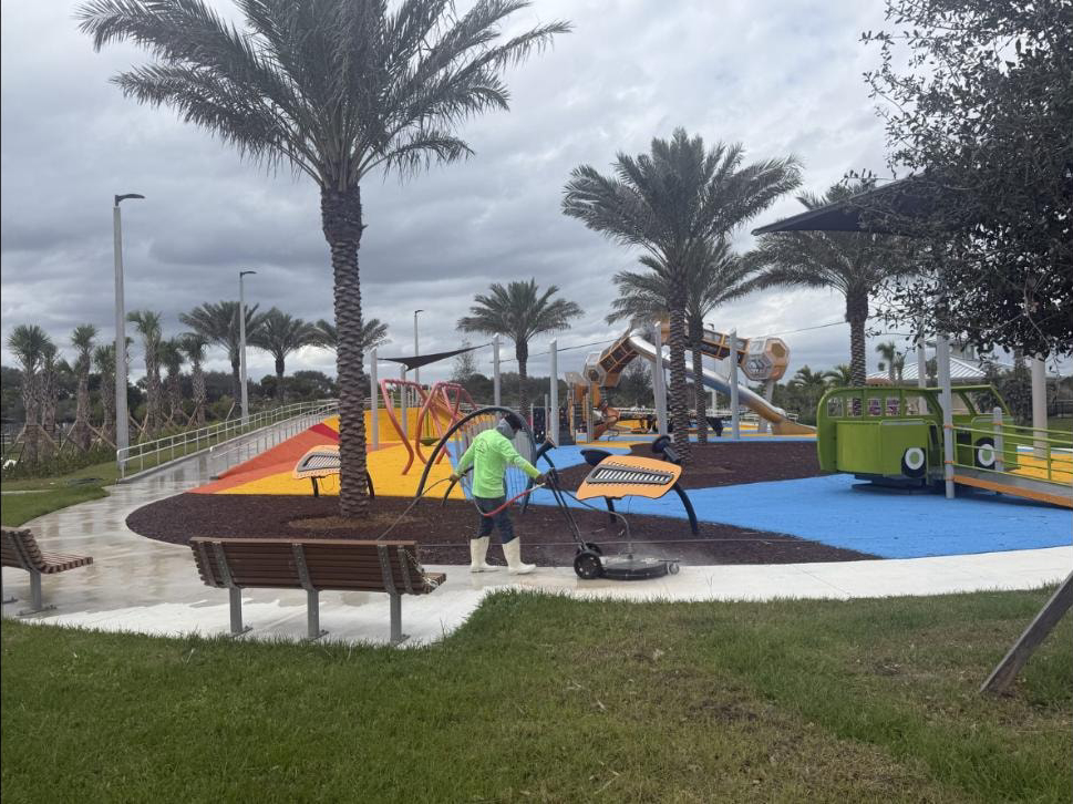 Technician pressure washing playground surfacing and surrounding concrete walkways at Canyon Park, Boynton Beach, FL — colorful rubber play surface, palm trees, overcast sky