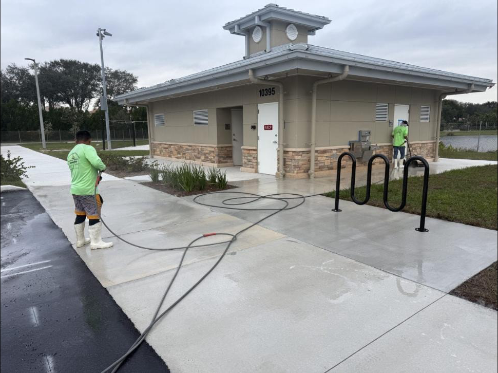 Two-man crew pressure washing the concrete walkway around a park restroom building at Canyon Park, Boynton Beach, FL — bike rack and overcast sky