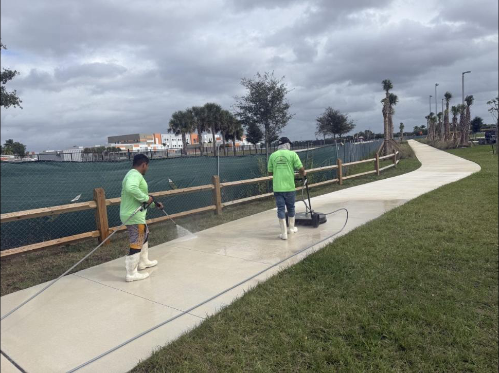 Two-man crew pressure washing a curved waterfront concrete walkway at Canyon Park, Boynton Beach, FL — surface cleaner and wand, lake and palm trees behind