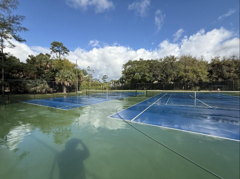 Tennis and pickleball courts freshly pressure washed in Lake Worth, FL — wet green and blue surface, hose across courts, tree canopy behind