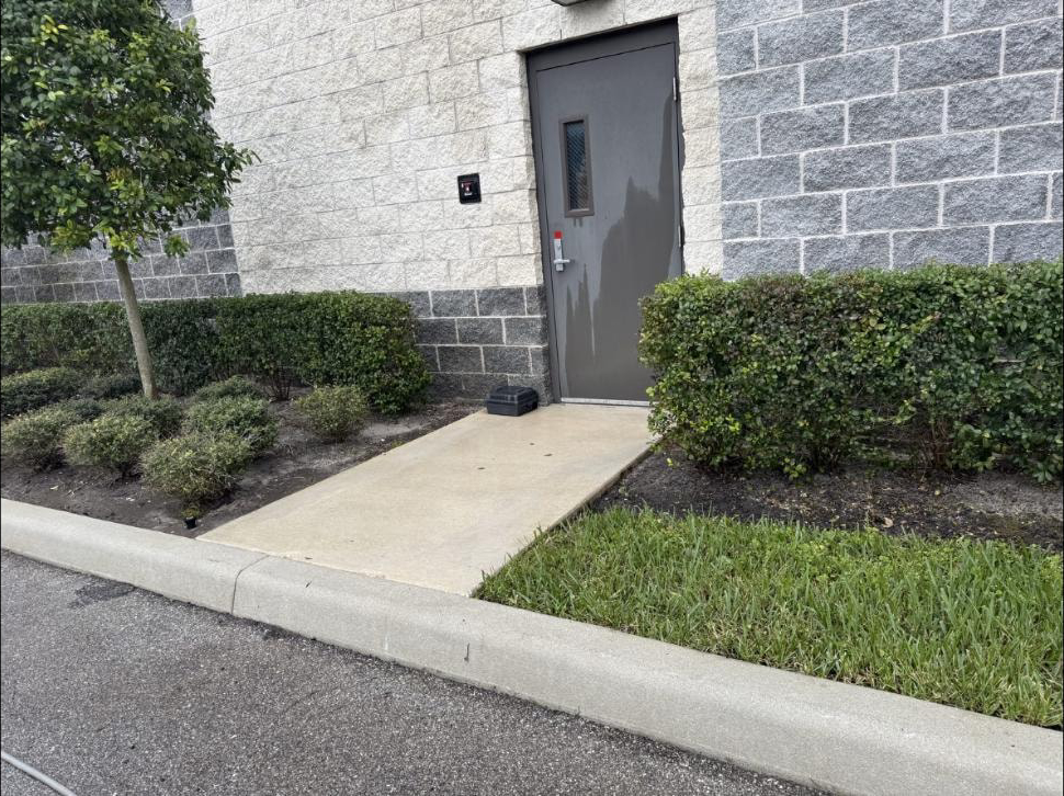 Freshly pressure washed concrete service walkway beside a commercial building in Palm Springs, FL — clean tan concrete, trimmed hedges, grey block wall exterior