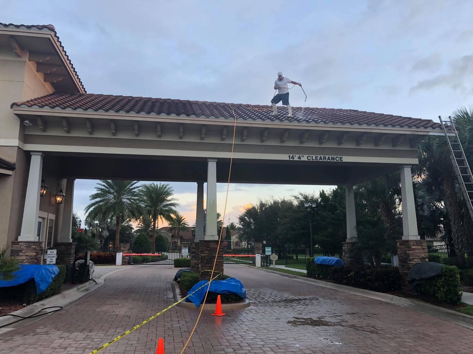 Before: HOA gatehouse roof during soft-wash cleaning, technician working on the barrel tile roof, staining still visible, Wellington, FL